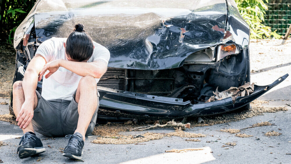 crying and desperate man sitting in front of his destroyed car after a vehicle collision